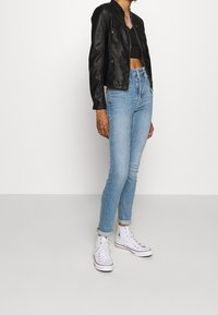 Giubbotto in pelle nera da biker, top corto aderente scuro, jeans skinny blu chiaro e sneaker alte bianche, che mettono in mostra la texture del denim e un design elegante.