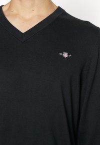 Pulover negru cu decolteu în V, realizat dintr-un material ribat, având un mic logo brodat pe partea stângă a pieptului, cu un design ajustat și o textură subtilă.