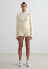 Krémový rebrovaný pletený top so zipsom a volánovými okrajmi, skombinovaný s rovnako tvarovanými šortkami. Model nosí biele športové tenisky.
