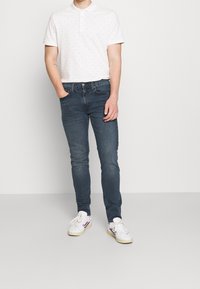 Vit polo med små färgglada prickar, matchad med mörkblå slim-fit jeans och vita sneakers med röda och blå detaljer.