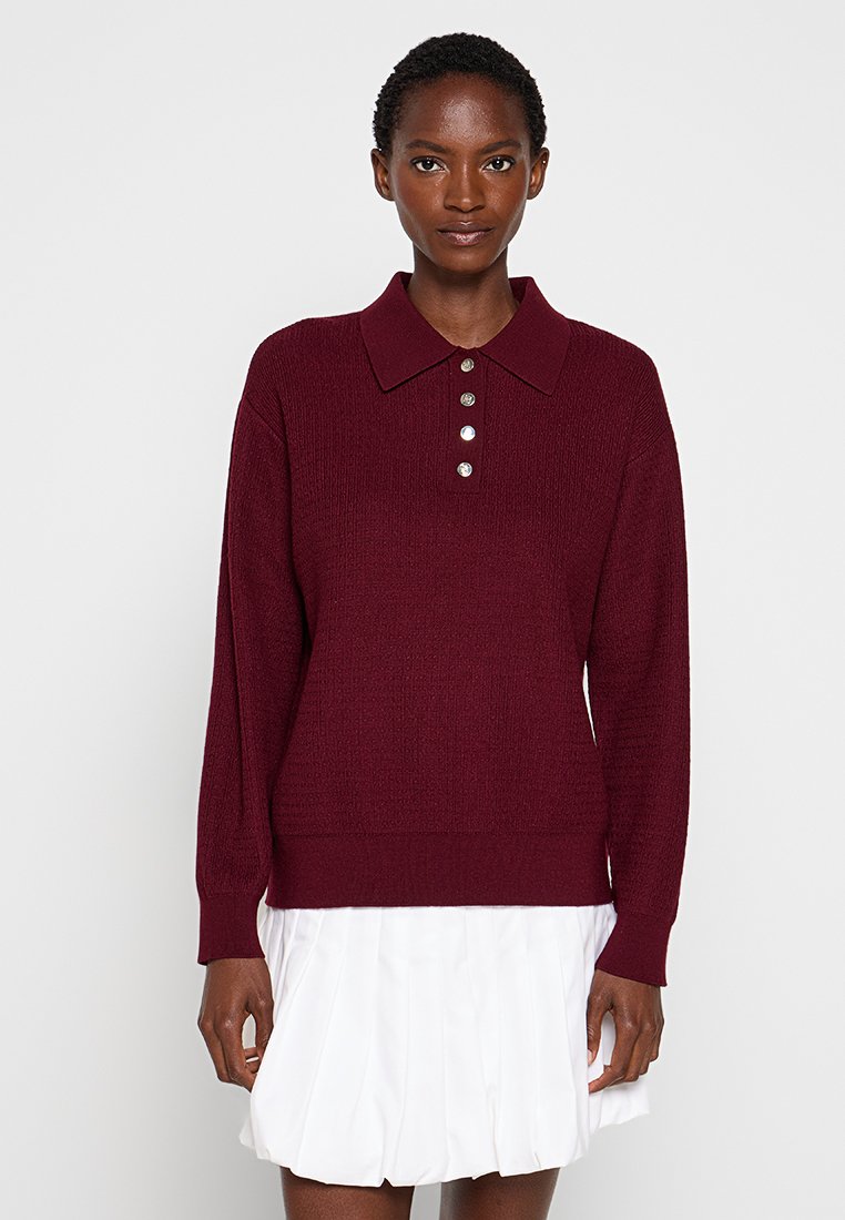 Pullover in magenta lavorato a maglia con colletto, cinque bottoni decorativi, maniche lunghe e orlo aderente. La texture è a coste e accogliente.