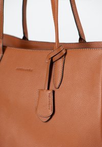 Tote-Bag aus tanfarbenem Leder mit strukturierter Oberfläche, zwei stabilen Griffen und einem seitlichen Akzentanhänger mit prägierter Markenbezeichnung. Sichtbare schwarze Kantenakzente.