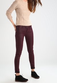 Beige Strickpullover mit strukturiertem Muster, kombiniert mit schmal geschnittenen burgunderfarbenen Jeans und schwarzen Schnürstiefeletten.
