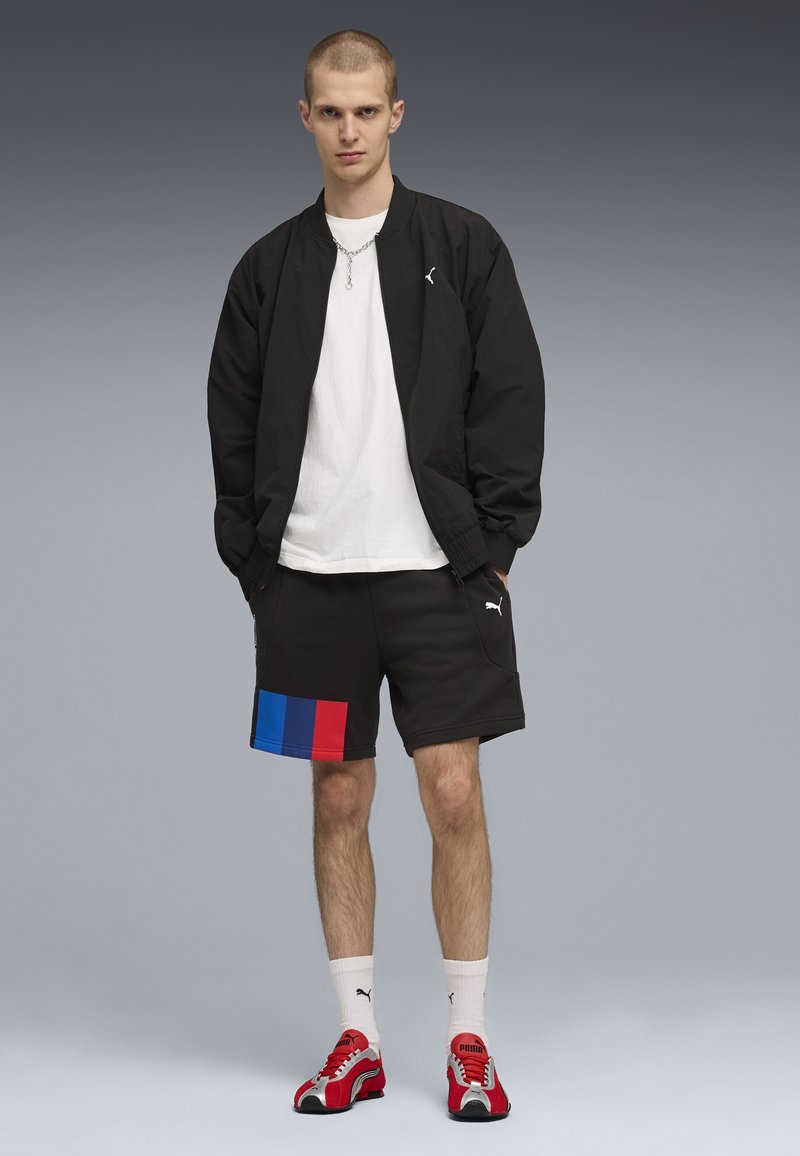 Blouson aviateur noir, t-shirt blanc, short noir avec blocs bleus et rouges, baskets rouges et chaussettes blanches. Le modèle est face à l'avant.