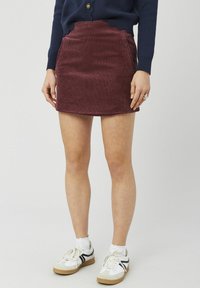 Mini-jupe en velours côtelé bordeaux avec une ceinture à festons, des poches latérales et une texture brossée, portée avec des baskets blanches.