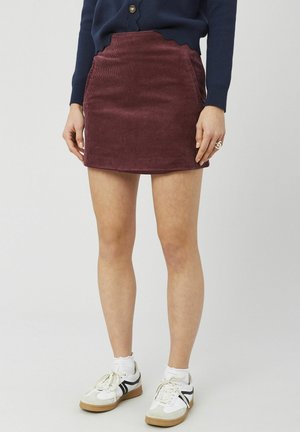 Mini-jupe en velours côtelé bordeaux avec une ceinture à festons, des poches latérales et une texture brossée, portée avec des baskets blanches.