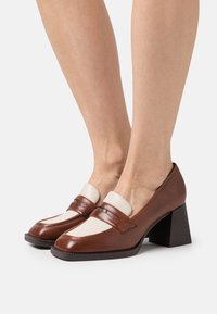 Zapatos de cuero marrón con tacón cuadrado, con un panel de color crema y costuras detalladas. Tienen una punta redondeada y acentos de hardware mínimos.