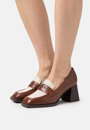Brown leather block-heeled shoes with a cream-colored panel and detailed stitching. Features a rounded toe and minimal hardware accents.