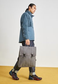 Sac à dos en cuir gris avec deux bretelles. Veste bleue doublée en polaire, pantalon sombre et chaussures noires avec des accents rouges et bleus.
