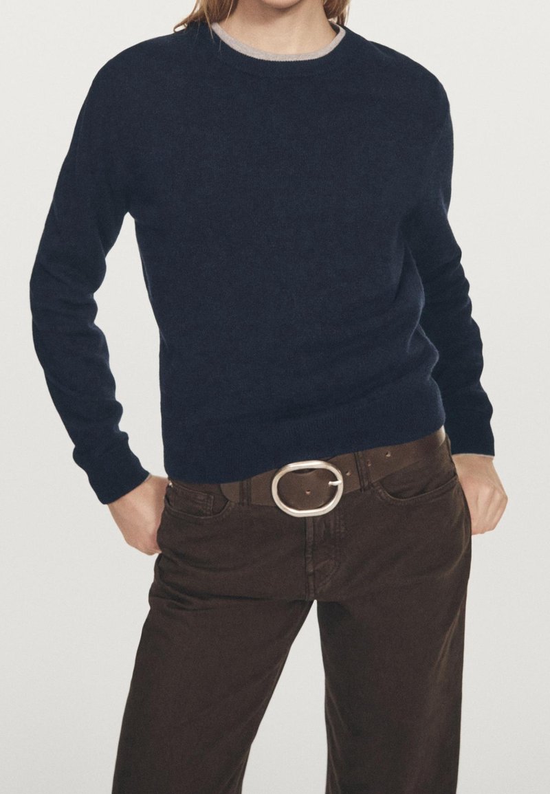 Personne portant un pull bleu marine foncé avec des bordures claires, un pantalon marron et une ceinture marron avec une grande boucle ovale en argent, les mains reposant près des poches.