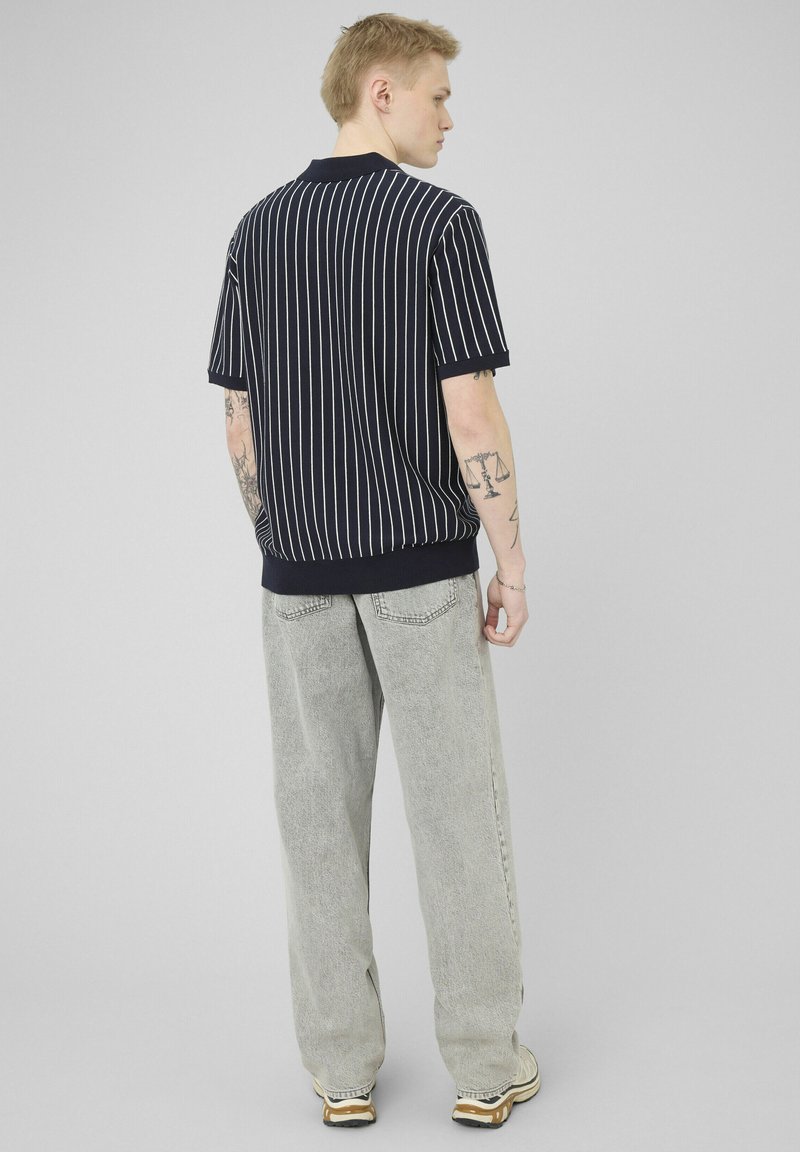 Jeune homme tatoué portant un t-shirt à manches courtes rayé noir et blanc et un jean ample gris clair, debout de dos.