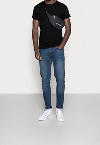 Czarna koszulka, niebieskie jeansy slim-fit, białe sneakersy oraz czarna nylonowa torba na ramię z zamkiem i detalem z logo. Jednolity tle.