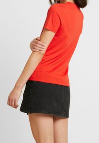 T-shirt in cotone rosso con maniche corte, design aderente e scollatura rotonda, indossato con una gonna di denim nera. Tessuto semplice, senza motivi visibili.