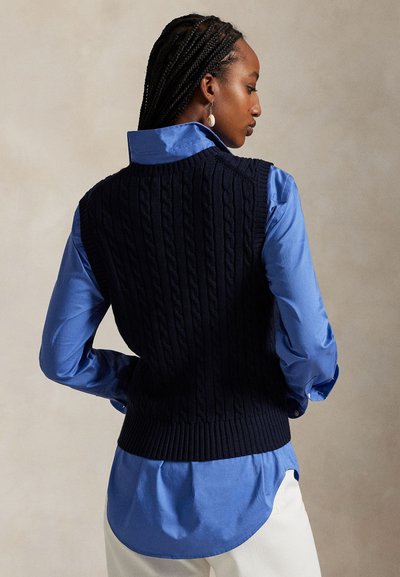 Μπλε σκούφο γιλέκο πλεκτό πάνω από ανοιχτό μπλε πουκάμισο, με ριμπ τελείωμα και γυρισμένο γιακά, συνδυασμένο με κρεμ παντελόνια, φαίνεται από πίσω.