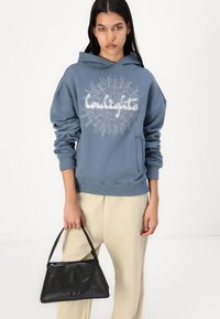 Sweatshirt azul com capuz e um design gráfico branco centrado no peito. Combinado com calças bege e uma bolsa preta texturizada.