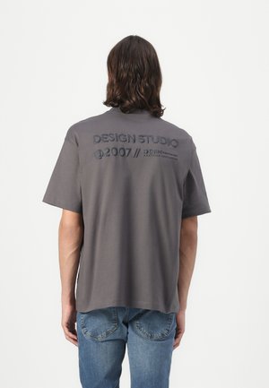 Gray cotton t-shirt with short sleeves, featuring raised text on the back: "DESIGN STUDIO @2007 // DENIM." Worn with blue jeans.