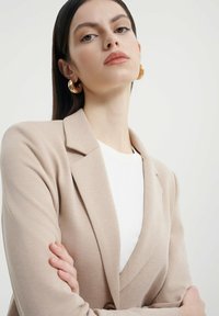 Beige sako se strukturovaným límcem, přiléhavým střihem a strukturovanou tkaninou. Nosené přes bílý top, s velkými zlatými kruhovými náušnicemi.