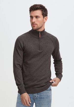 Homme portant un pull gris foncé à demi-fermeture éclair et un jean bleu clair, regardant vers sa gauche sur un fond blanc uni.