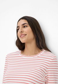 Weißes Langarmoberteil mit roten horizontalen Streifen, rundem Halsausschnitt und glatter Stofftextur. Dezente Nahtdetails am Halsausschnitt.