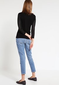 Schwarzes Oberteil mit gestreiften langen Ärmeln, kombiniert mit hellblauen Jeans. Das Modell trägt schwarze Ballerinas und wird von hinten betrachtet.