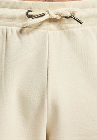Gros plan sur la ceinture d'un pantalon de survêtement beige avec un élastique côtelé et un cordon noué avec embouts métalliques.