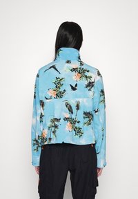 Person with long black hair wearing a light blue jacket with floral and bird patterns, paired with black cargo pants, facing away.