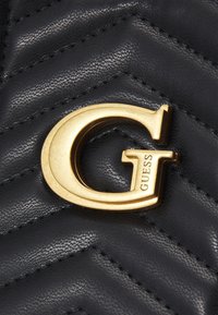 Złote metalowe logo "G" z wygrawerowanym napisem "GUESS" na czarnej, pikowanej skórze z diagonalnym przeszyciem.