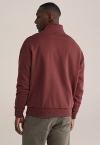 Bordová mikina s vysokým golierom a rebrovanými manžetami. Mäkká, textúrovaná tkanina; voľný strih. Má viditeľný šev a minimalistický dizajn.