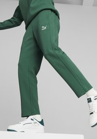 Grüne Sporthose mit glatter Textur, auf dem Oberschenkel mit dem weißen Puma-Logo versehen. Kombiniert mit weißen Sneakers und kontrastierenden tealfarbenen Akzenten.