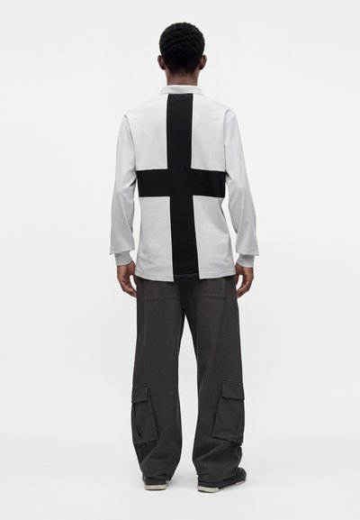 Homme de dos portant une chemise blanche à manches longues avec une grande croix noire dans le dos, un pantalon cargo foncé et des baskets noires.