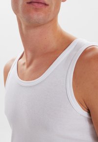 Hvid bomulds tanktop med ribbet tekstur, der har en bred halsudskæring og tynde stropper. Beklædningen er figursyet og uden sømme i kanterne.