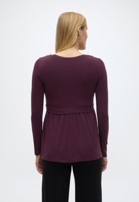 Γυναίκα με ίσια ξανθά μαλλιά, που φοράει μακρυμάνικη σκούρα μοβ μπλούζα και μαύρο παντελόνι, γυρισμένη προς τα πίσω σε απλό ανοιχτόχρωμο φόντο.