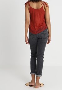 Rode suède tanktop met kanten rand aan de zoom, gecombineerd met donkergrijze opgerolde jeans en platte sandalen, gezien vanaf de achterkant.