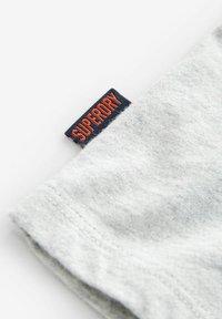 Grå bomullstyg med en mjuk textur, med en blå och orange "SUPERDRY" märkesetikett vid fållen. Tydlig sömdetalj synlig.
