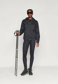 Veste légère noire avec capuche, associée à des leggings noirs présentant différentes textures. Tient un bâton de randonnée, porte des lunettes de soleil foncées et des bottes.