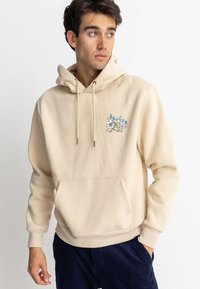 Sudadera con capucha beige con un bolsillo frontal y un gráfico decorativo de flores azules. Confeccionada con tela suave, cuenta con cierre de cordón y puños acanalados.