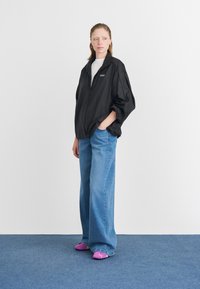 Ung kvinde står på blå tæppe iført en oversized sort jakke, hvid skjorte, brede blå jeans og lilla Puma sneakers, mens hun kigger til siden.