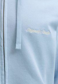 Φωτεινό γαλάζιο φούτερ με φερμουάρ, με υφασμένο κορδόνι και μικρό λευκό κεντητό κείμενο "Originals Studio" στο στήθος.