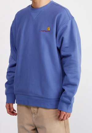Homme portant un sweat-shirt Carhartt bleu à col rond à manches longues et un pantalon beige sur un fond uni.