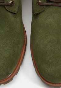 Primo piano di scarpe in suede verde con suole in gomma marrone e lacci marrone scuro, che mostrano i dettagli della texture e delle cuciture.