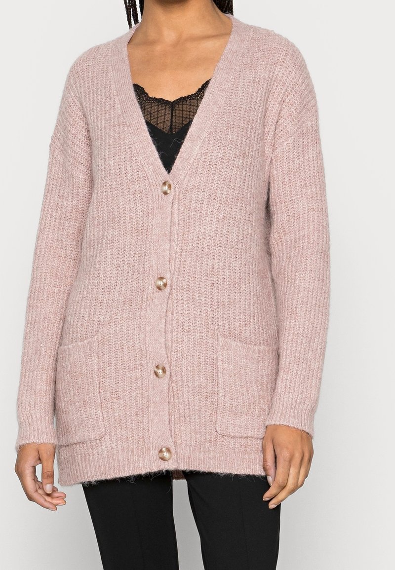 Femme portant un cardigan en tricot rose pâle avec de gros boutons par-dessus un top en dentelle noire et un pantalon noir, debout devant un fond uni.
