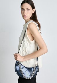 Torba typu hobo z tie-dye w odcieniach niebieskiego i białego, z akcentem srebrnego łańcucha i czarnym paskiem, zestawiona z kremową, dzianą kamizelką.