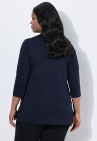 Frau mit langen, welligen Haaren, die ein navyblaues Dreiviertelarmhemd trägt, steht vor einem weißen Hintergrund und schaut zur Seite.