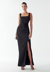 Žena stojící v bezramínkových černých večerních šatech s rozparkem až ke stehně, držící psaníčko a obutá v sandálech na vysokém podpatku a zlatých špercích.