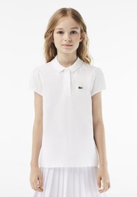 Biała koszulka polo z krótkimi bufkowymi rękawami, kołnierzykiem i zielonym haftowanym logo na lewej piersi. Miękka bawełniana struktura, klasyczny krój.
