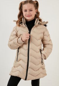 Beige dunjakke med chevron-mønster, lynlås foran, hætte og sorte accenter. Har elastiske manchetter og sidelommer.