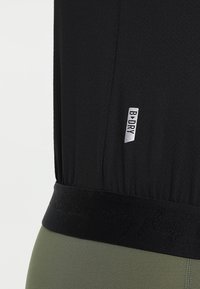 Top atlético negro con tejido texturizado, que presenta una prominent etiqueta B:DRY. El dobladillo tiene una banda elástica negra con un sutil detalle de texto.