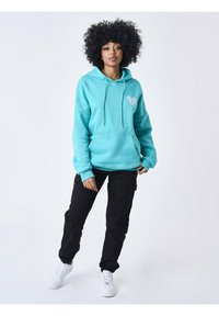 Sudadera azul claro con un gráfico de un corazón en el lado izquierdo del pecho, combinada con pantalones cargo negros y zapatillas blancas. Tejido suave, corte holgado.