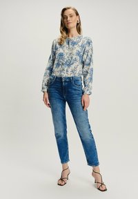 Kobieta w niebieskiej kwiatowej bluzce z długim rękawem, w niebieskich spodniach z wysokim stanem i czarnych sandałach na obcasie z paskami, stoi na jednolitym tle.