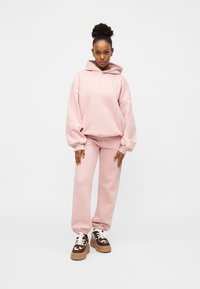 Sweat à capuche oversize rose pâle avec cordon de serrage et joggeurs assortis à coupe fuselée. Porté avec des baskets marron à motifs et semelles épaisses.